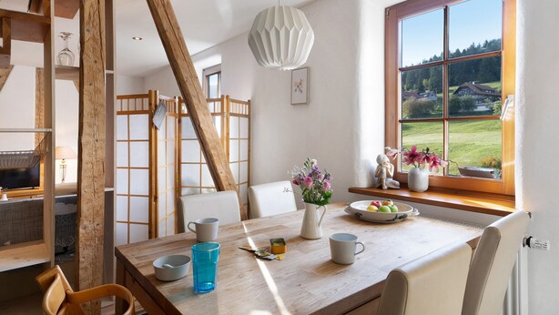 A dining area with a wooden table, chairs, a vase with flowers, and a view of a green landscape through a window. - Apartment 'Familienwohnung Nala' with Mountain View, Shared Garden and Wi-Fi (Forbach)