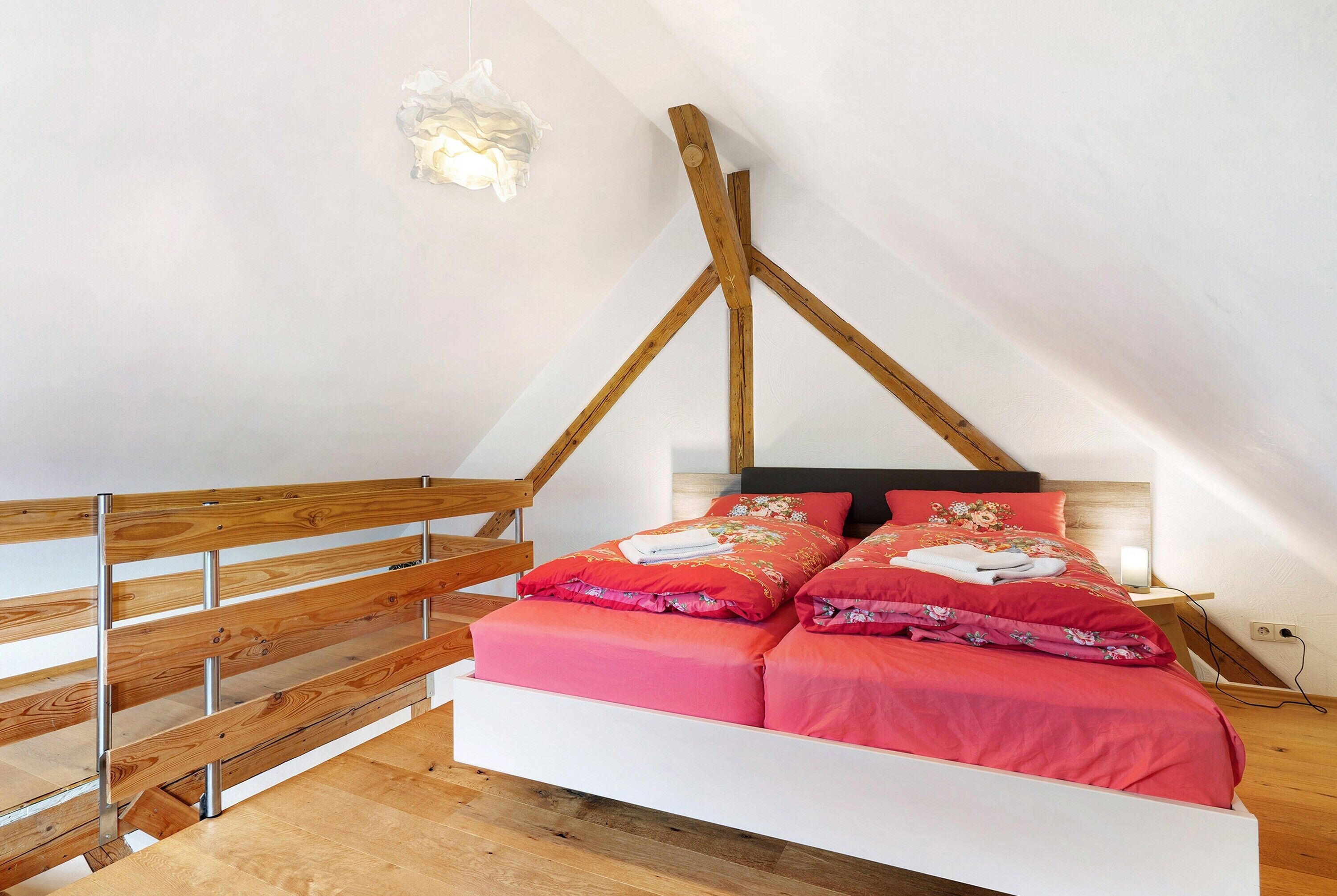 Quarto com teto inclinado, vigas de madeira, cama branca com roupa de cama vermelha e corrimão de madeira.