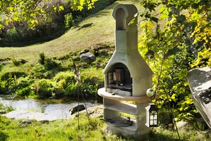 A stone-built outdoor oven with a chimney, surrounded by greenery and a small stream.