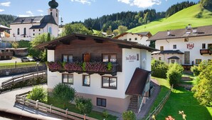 Un chalet traditionnel doté d’un balcon, une église en arrière-plan et une colline verdoyante.