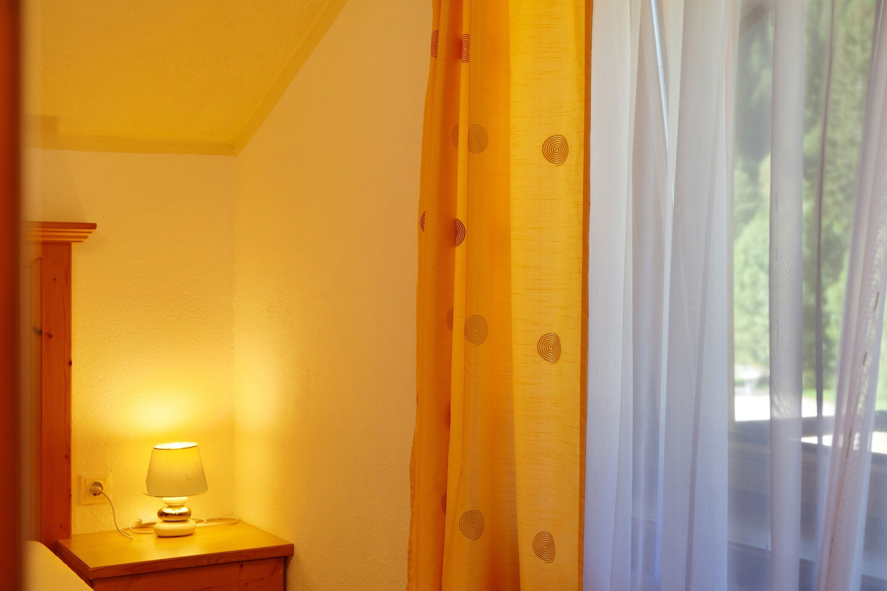 Een kamer met een bedlampje, een raam met dunne gordijnen en uitzicht op groen.