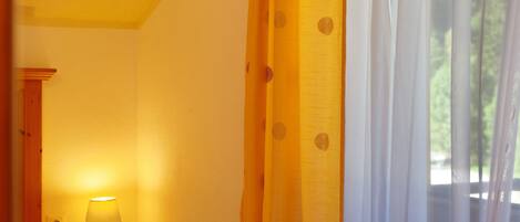 A room with a bedside lamp, a window with sheer curtains, and a view of greenery outside.