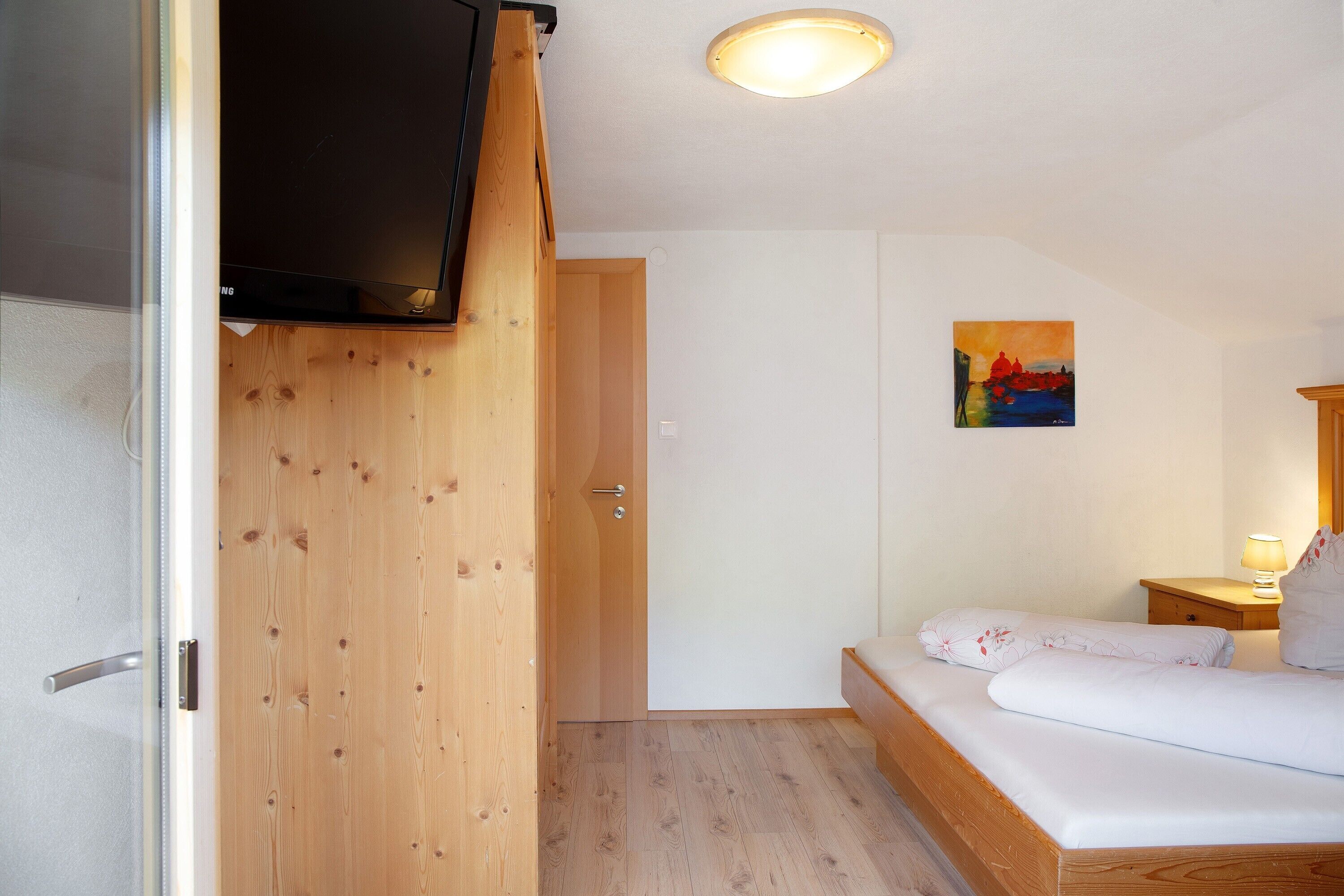 A bedroom with a bed, a wooden wardrobe, a television mounted on the wall, a painting, and a bedside table with a lamp.