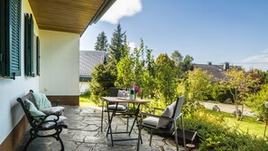 Une terrasse avec un sol en pierre, deux chaises, une table avec une bouteille et des verres, et une vue sur un jardin et les maisons voisines.