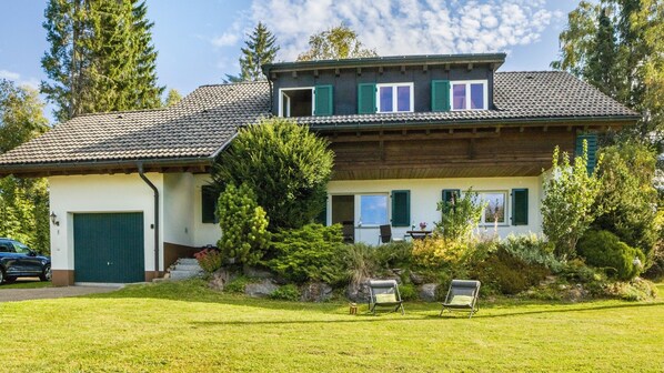Une maison à deux étages avec une porte verte, un porche abrité et une pelouse où sont disposées deux chaises.