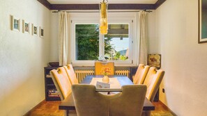 Une salle à manger avec une table, des chaises, un vase de fleurs, une fenêtre donnant sur un beau paysage et des tableaux encadrés accrochés au mur.