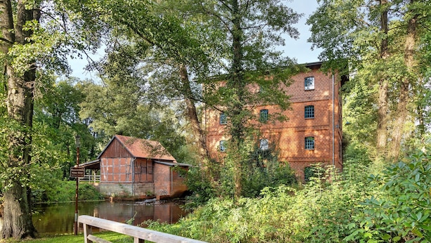 A wooden bridge leading to a brick building by a calm river, surrounded by lush greenery.