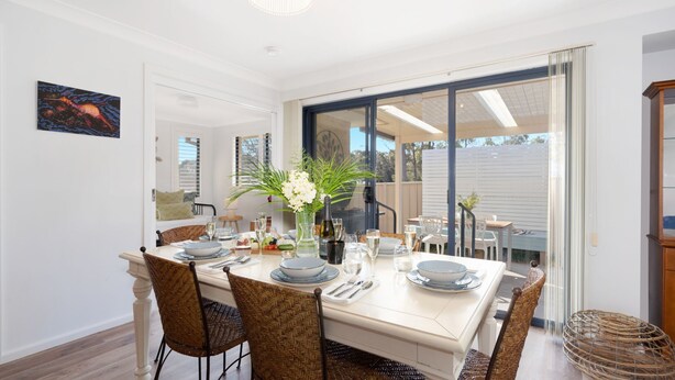 A dining area with a white table set for six, surrounded by wicker chairs, a vase of flowers, and a bottle of wine. - The Beach Bach – Peaceful and relaxing villa (Vincentia)