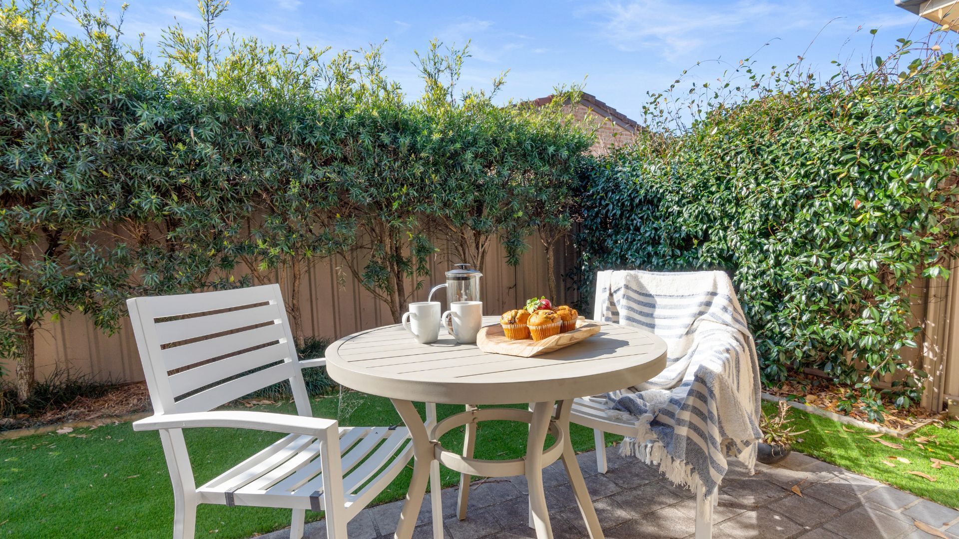 Une table ronde avec deux chaises blanches, une cafetière et un plateau de muffins.
