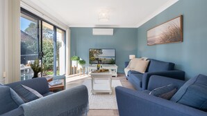 A modern living room with a large TV, blue sofas, and a view of the outdoors through large windows. - The Beach Bach – Peaceful and relaxing villa (Vincentia)