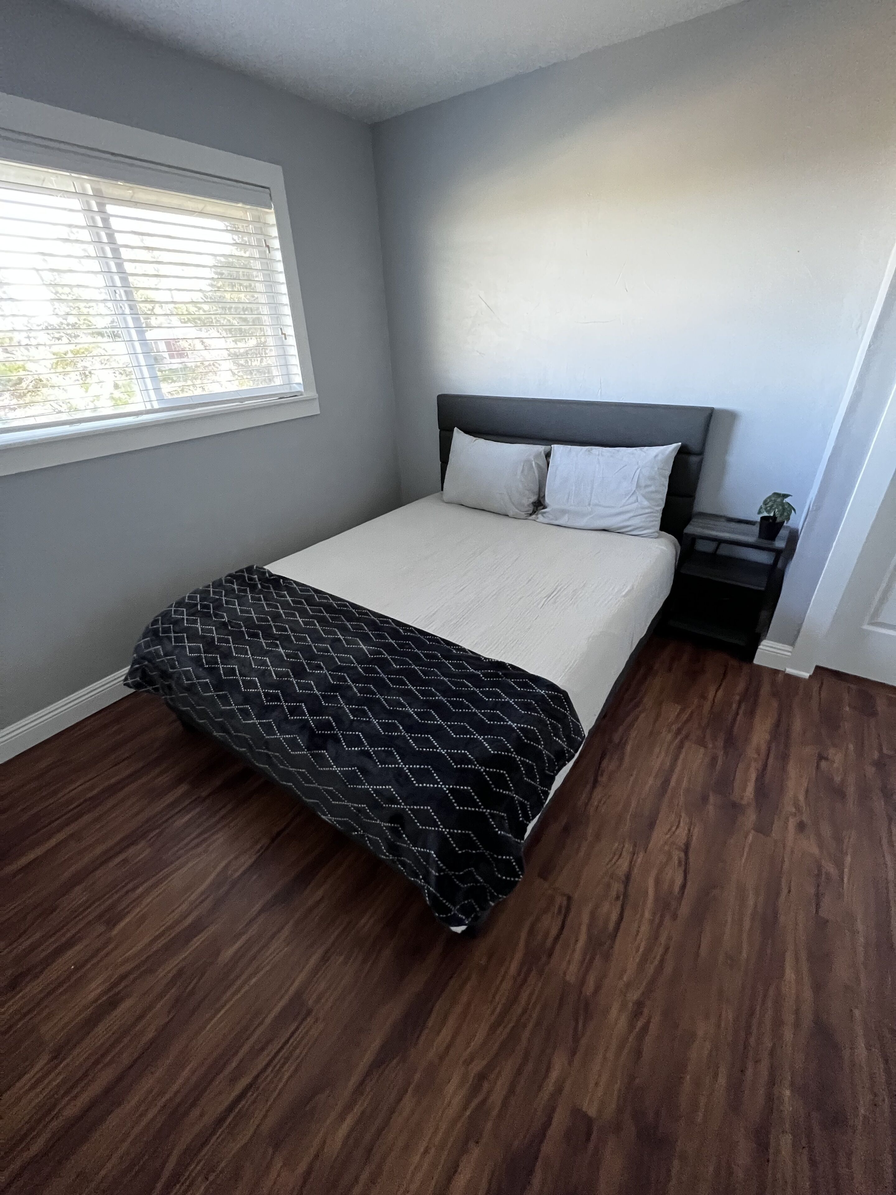 A bedroom with a bed, a nightstand, a window, and wooden flooring.