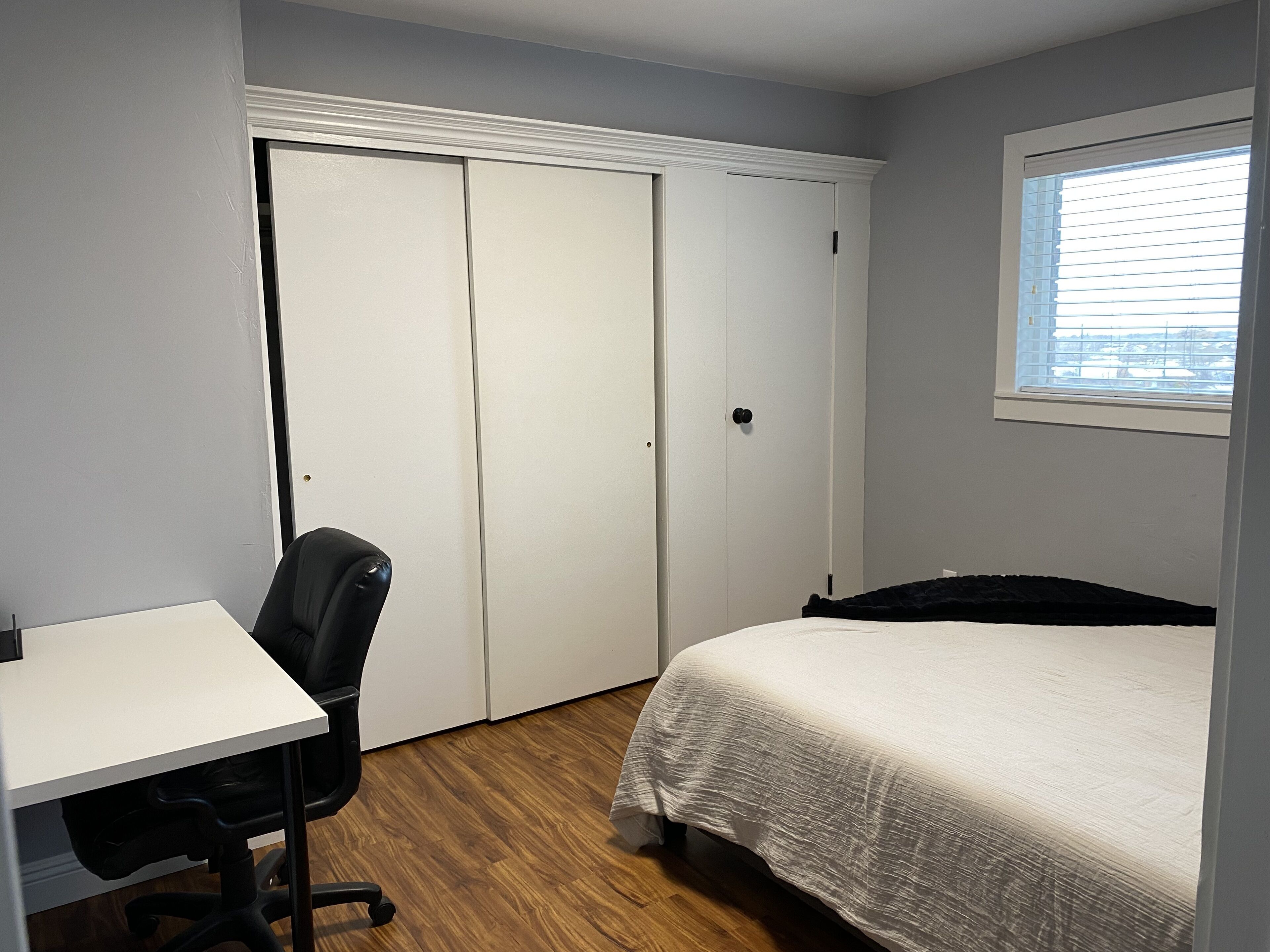 A bedroom with a bed, a desk, and a chair. There is a window with blinds and a door with a handle.