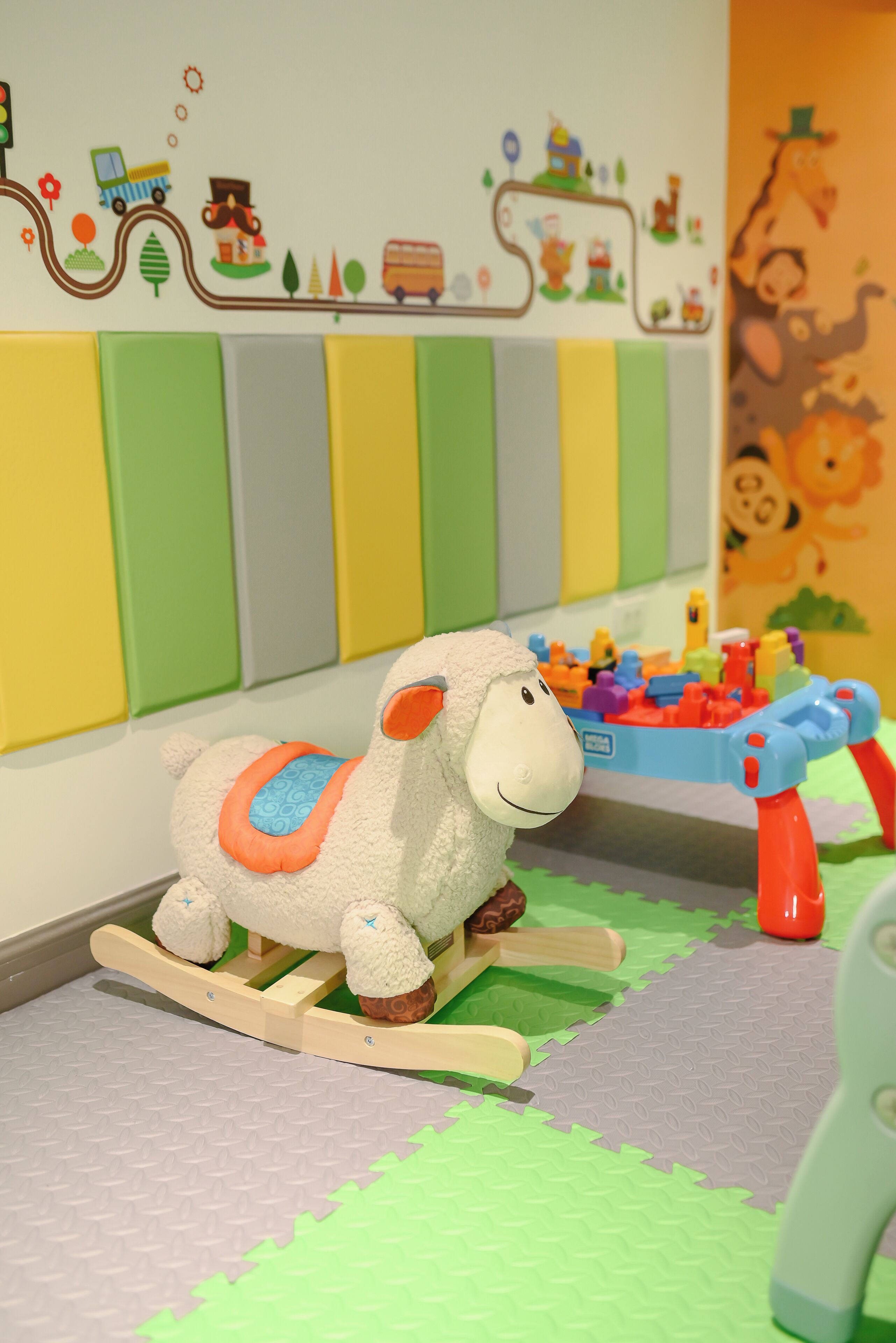 A toy sheep on a rocking horse, a toy table with colorful blocks, and a wall with a colorful mural.