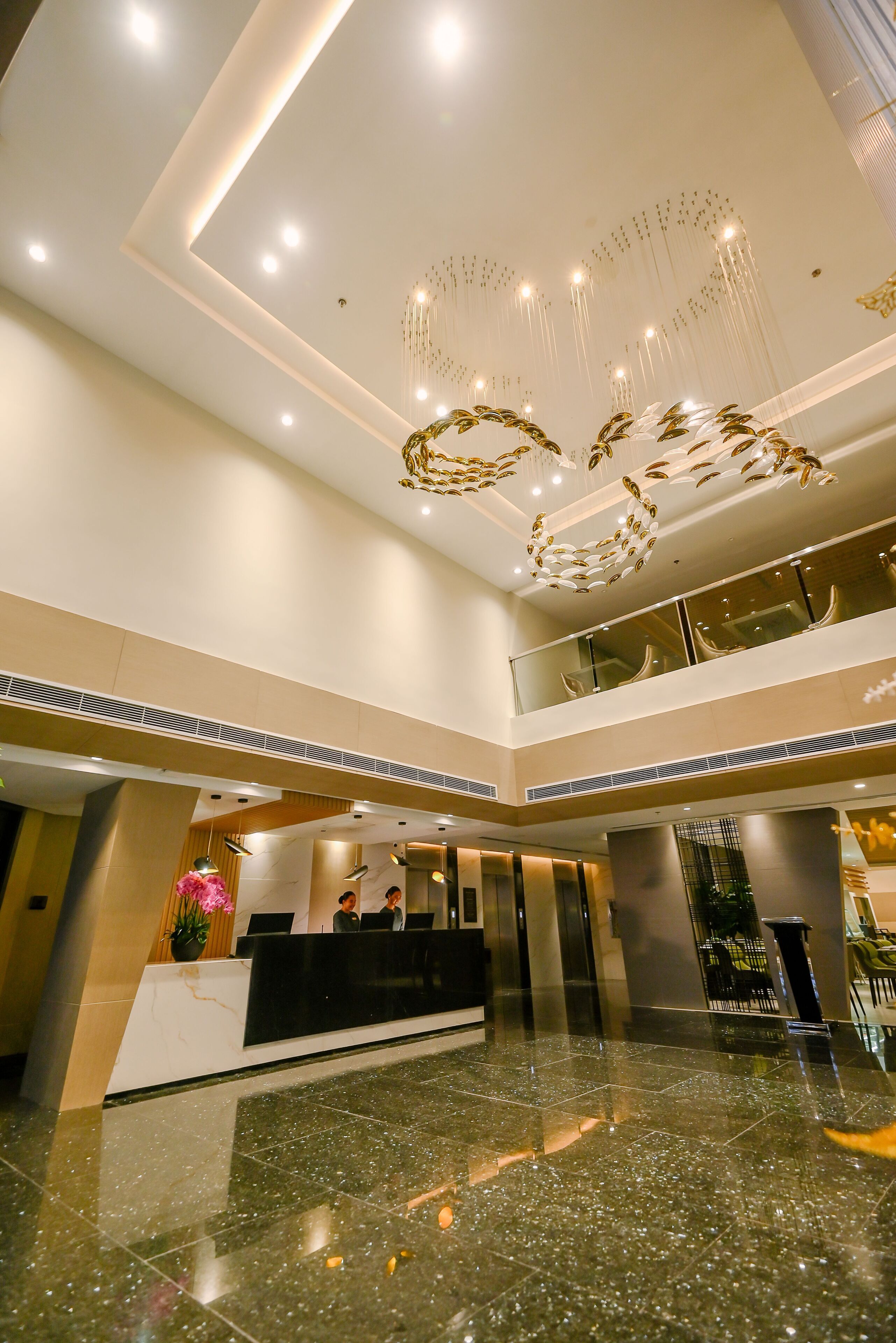 Un moderno vestíbulo de hotel con una recepción, baranda de vidrio en un nivel superior y una lámpara decorativa en el techo.