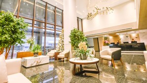 Um lobby moderno com balcão de recepção em mármore, assentos confortáveis, janelas amplas e plantas decorativas.