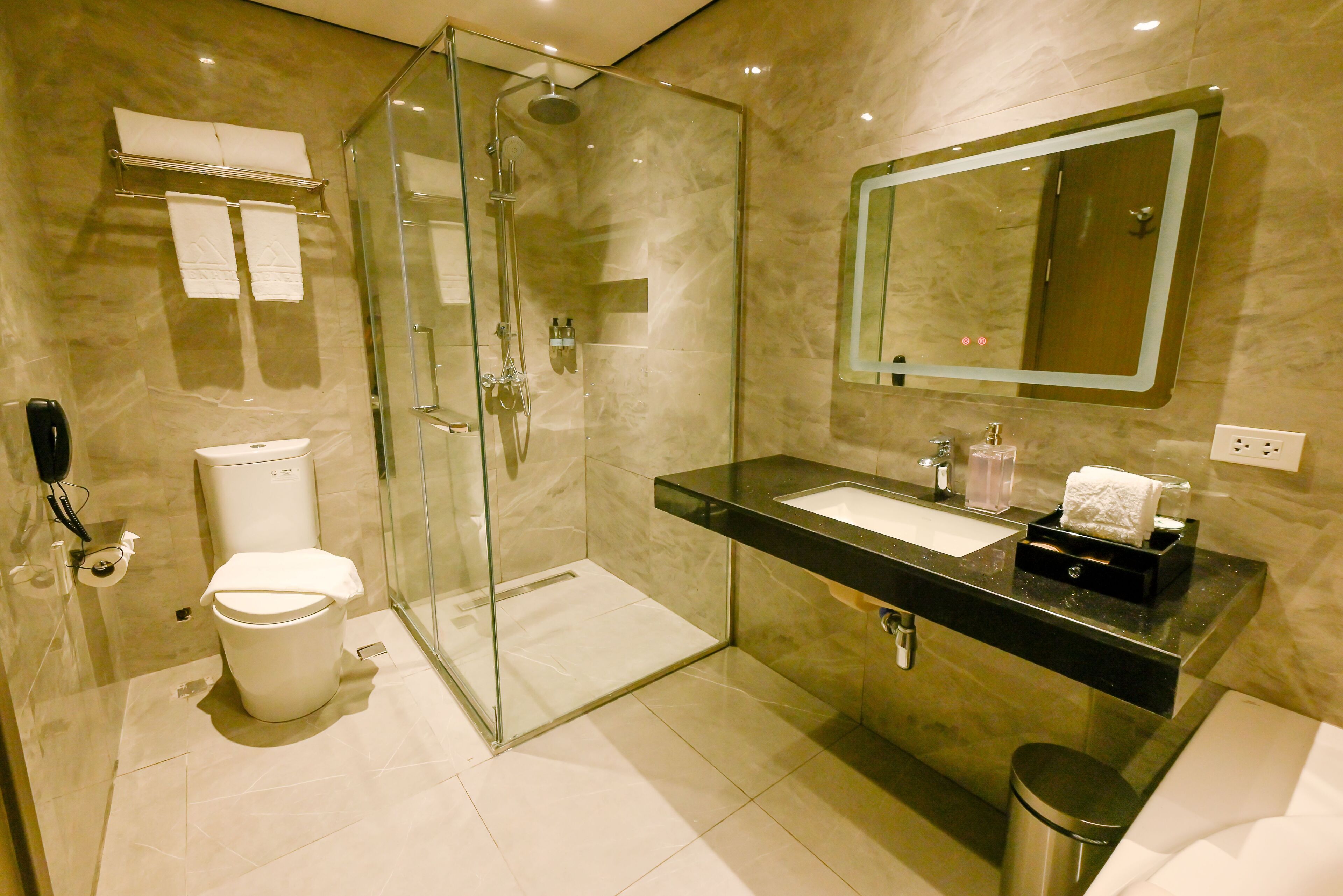 Une salle de bain moderne avec une douche à parois de verre, des toilettes blanches, un lavabo noir et un miroir au-dessus du lavabo.