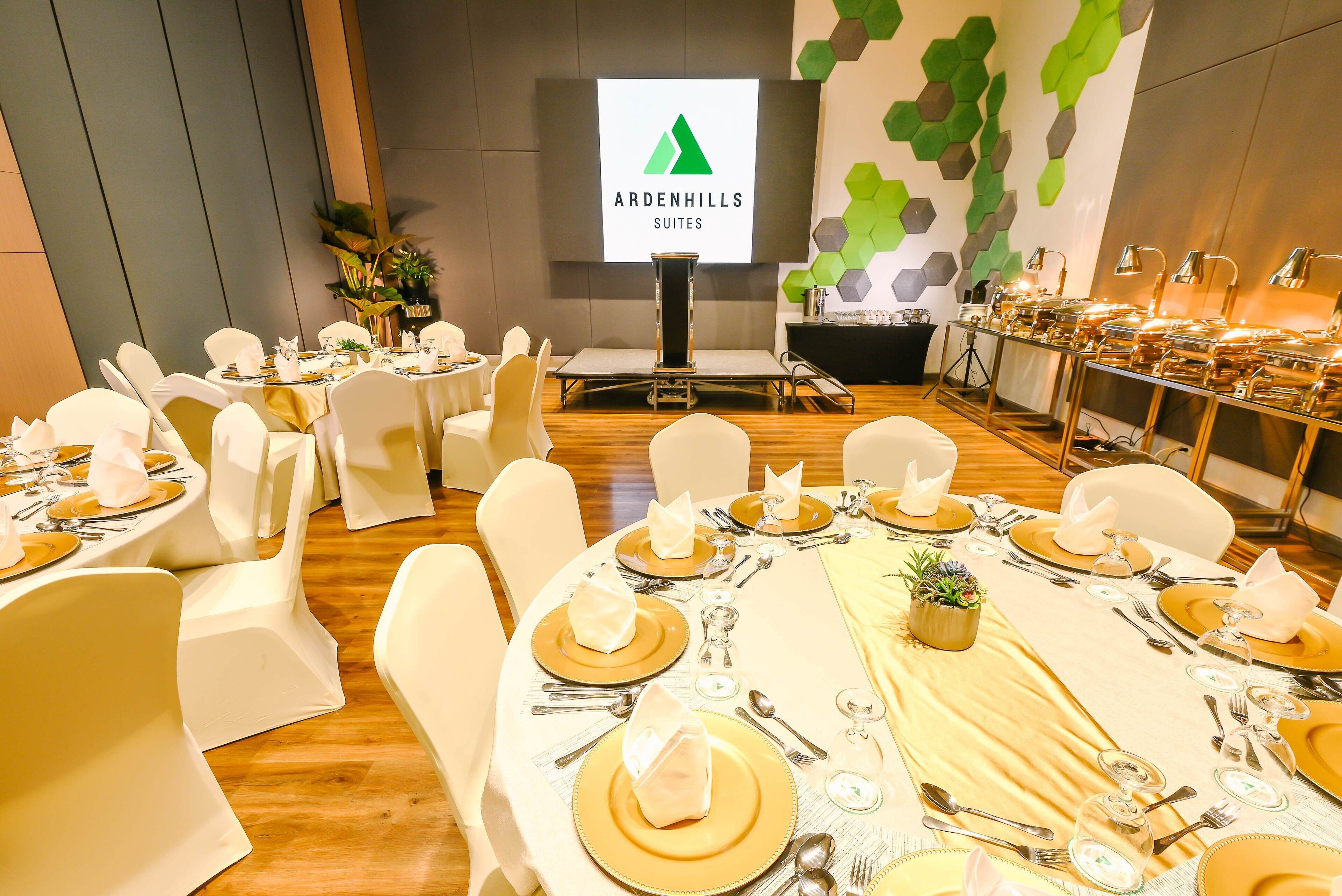 Une salle de conférence avec des tables disposées pour un événement formel, un écran de projection affichant le texte « Ardenhills Suites », et un design mural moderne.