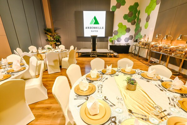 Un salón de conferencias con mesas dispuestas para un evento formal, una pantalla de proyección que muestra 'Ardenhills Suites' y un diseño de pared moderno.