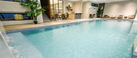An indoor swimming pool with clear water, surrounded by a modern interior with a seating area, a staircase, and a kitchen counter.