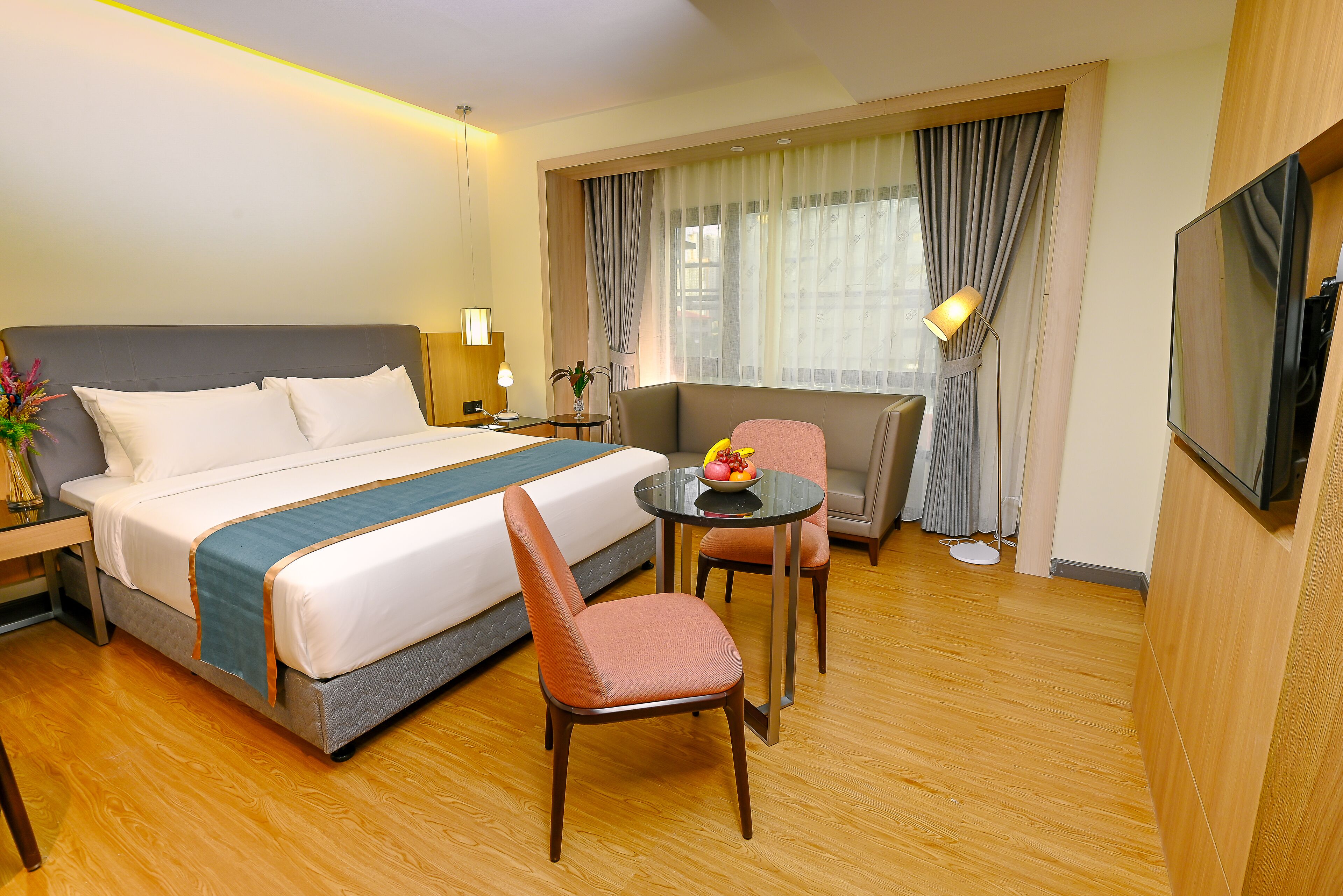 Quarto de hotel com cama, escrivaninha, cadeira, mesa pequena com abajur e televisão.