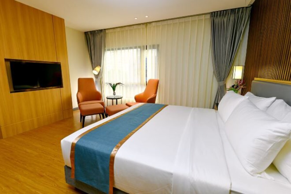 A hotel room with a large bed, two armchairs, a small table, a TV mounted on the wall, and a window with curtains.