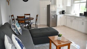 Una sala de estar moderna con un sofá gris, sillas de madera, una mesita de madera y una zona de cocina con armarios blancos y un refrigerador.
