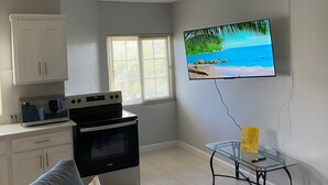 Una sala de estar con un sofá gris, un televisor montado en la pared, un ventilador de techo y una cocina con armarios y electrodomésticos.