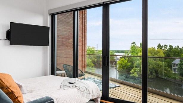 A bedroom with a bed, a TV mounted on the wall, and a large sliding glass door leading to a balcony with a view of greenery and a river. - Pine Tree House 316 (Traverse City)