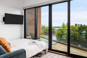 A bedroom with a bed, a TV mounted on the wall, and a large sliding glass door leading to a balcony with a view of greenery and a river.