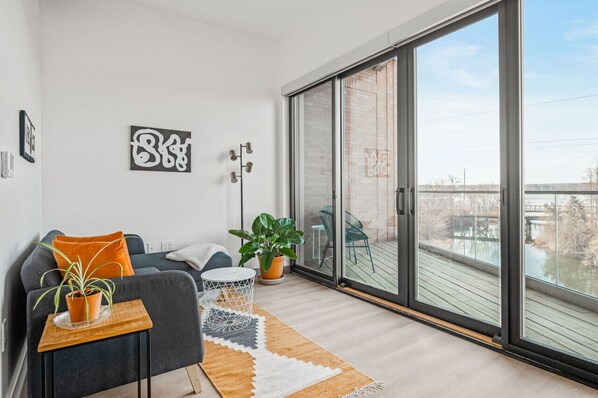 A modern living room with a grey sofa, a small table with a plant, and a large sliding glass door leading to a balcony with a view of a bridge and trees.