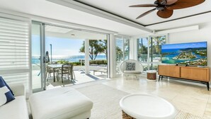 A modern living room with a large TV, a ceiling fan, and a view of the ocean.