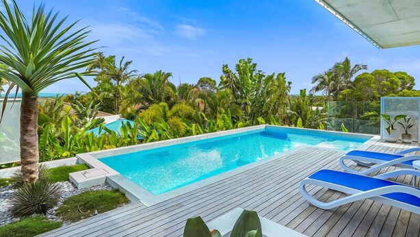 Piscina com espreguiçadeiras, rodeada por plantas tropicais e um deck de madeira.