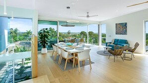 Uma sala de estar ampla com mesa de jantar, cadeiras e sofá. Há uma grande janela com vista para vegetação e paisagem distante.