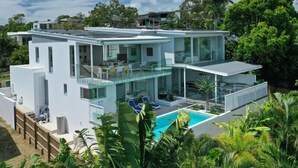 Uma casa moderna de dois andares com varanda de vidro, piscina e vegetação exuberante.