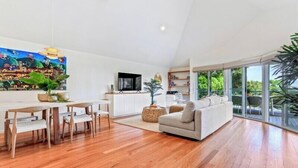A spacious living area with a wooden floor, a sofa, a dining table with chairs, a flat-screen TV, and a large window offering a view of greenery.