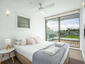 Quarto com cama grande, mesa de cabeceira, ventilador de teto, janela com vista para um jardim e porta de vidro deslizante que dá para uma área externa.