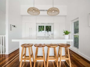 Uma cozinha com ilha branca, banquetas de madeira e duas luminárias pendentes de vime.