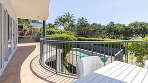 Un balcón con piscina, mobiliario exterior blanco y vistas a palmeras y follaje.