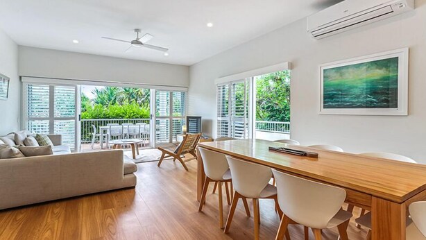 Amplia sala de estar con mesa de comedor de madera, sillas blancas, sofá y ventanas grandes con vista a la vegetación.