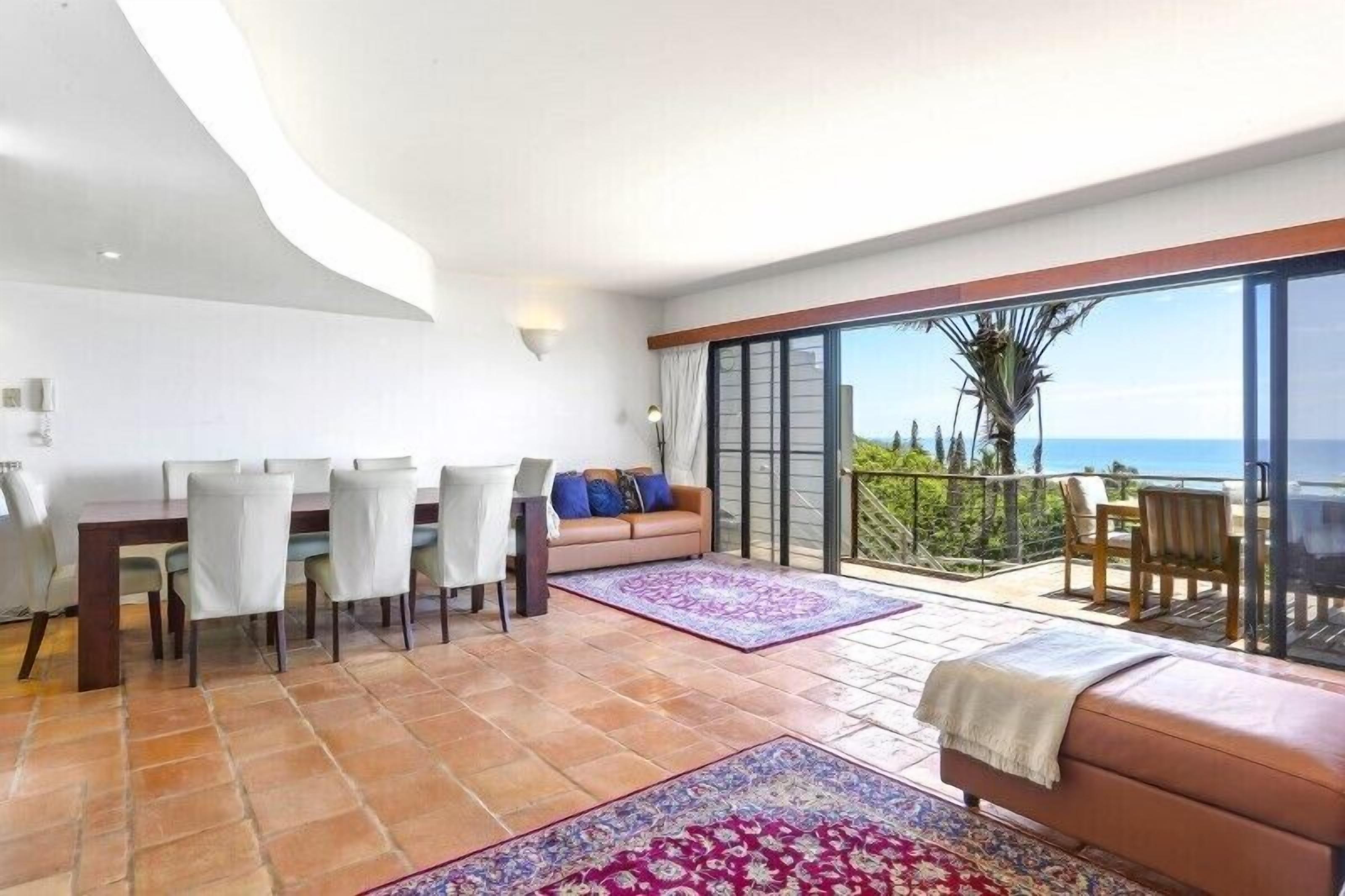 A spacious living area with a dining table, chairs, a sofa, and a balcony with a view of the sea.