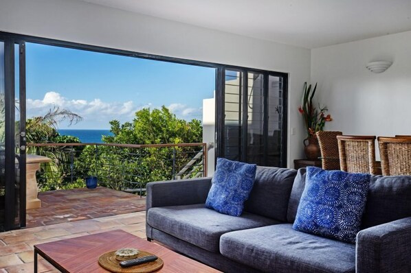 A modern living room with a grey sofa, two blue patterned cushions, a wooden coffee table, and a view of the ocean through large glass doors. - A Mediterranean Getaway, Sunshine Beach (Sunshine Beach)