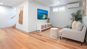 A modern living room with a wooden floor, a white sofa, a white cabinet, a round wicker side table, and a large painting of a beach scene. - Luxe Coastal Abode, Marcus Beach (Marcus Beach)