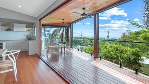 A modern kitchen with white cabinets, a dining area with a white chair, and a balcony with a view of greenery and a body of water. - Luxe Coastal Abode, Marcus Beach (Marcus Beach)