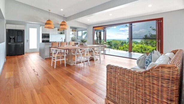 A spacious living area with a wooden floor, a dining table with chairs, a wicker sofa, and a large sliding glass door leading to a balcony with a view of greenery. - Luxe Coastal Abode, Marcus Beach (Marcus Beach)