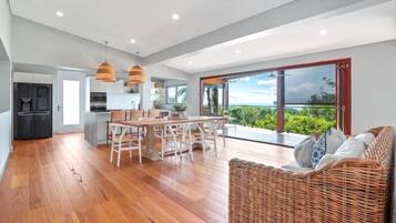 A spacious living area with a wooden floor, a dining table with chairs, a wicker sofa, and a large sliding glass door leading to a balcony with a view of greenery.