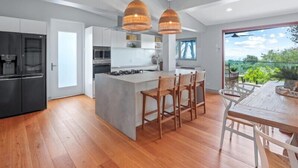A modern kitchen with a central island, wooden chairs, and pendant lighting. - Luxe Coastal Abode, Marcus Beach (Marcus Beach)