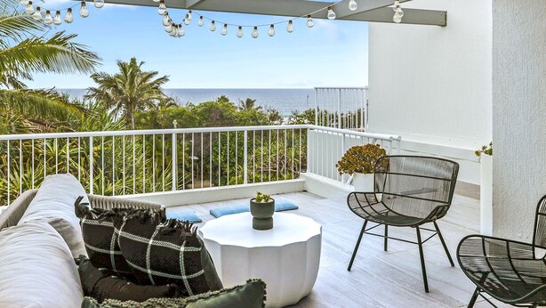 A balcony with a white railing, a round table, and two chairs. There is a string of lights hanging above and a view of the ocean and palm trees in the background.