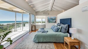 A bedroom with a large bed, a nightstand, a view of the ocean, and a ceiling with exposed beams. - Indulge at Iluka, Castaways Beach (Noosa)
