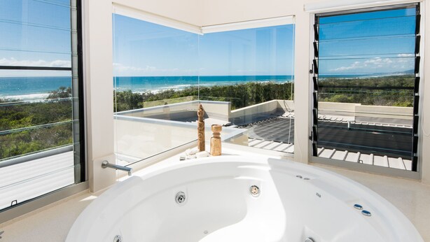 A modern bathroom with a large oval bathtub, large windows offering ocean views, and a balcony with a glass railing. - Indulge at Iluka, Castaways Beach (Noosa)
