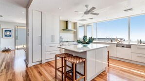 A modern kitchen with white cabinets, a central island with stools, a ceiling fan, and large windows offering a view. - Indulge at Iluka, Castaways Beach (Noosa)